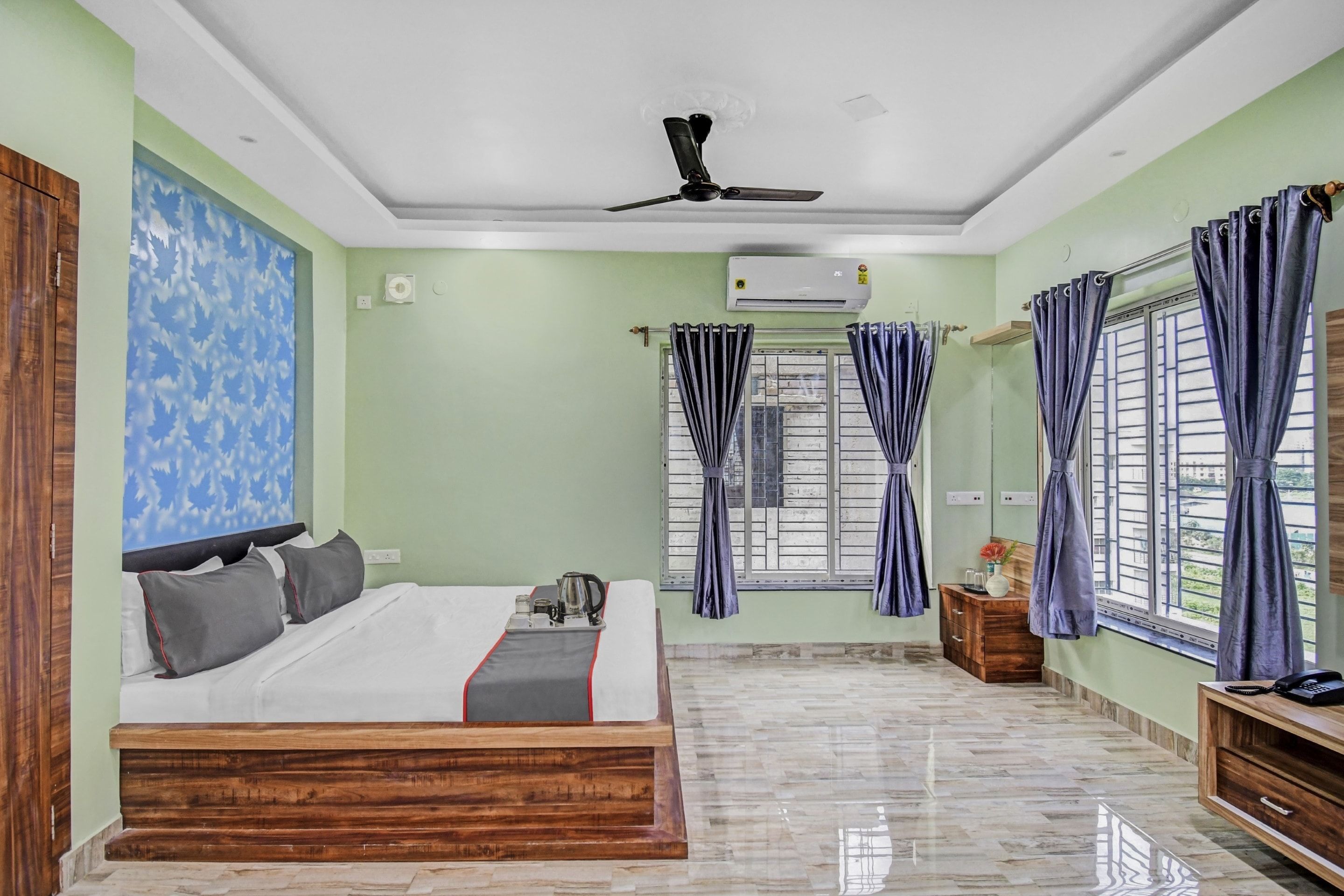 A hotel room with a bed, a ceiling fan, an air conditioning unit, a window with curtains, a wooden nightstand, and a marble floor.
