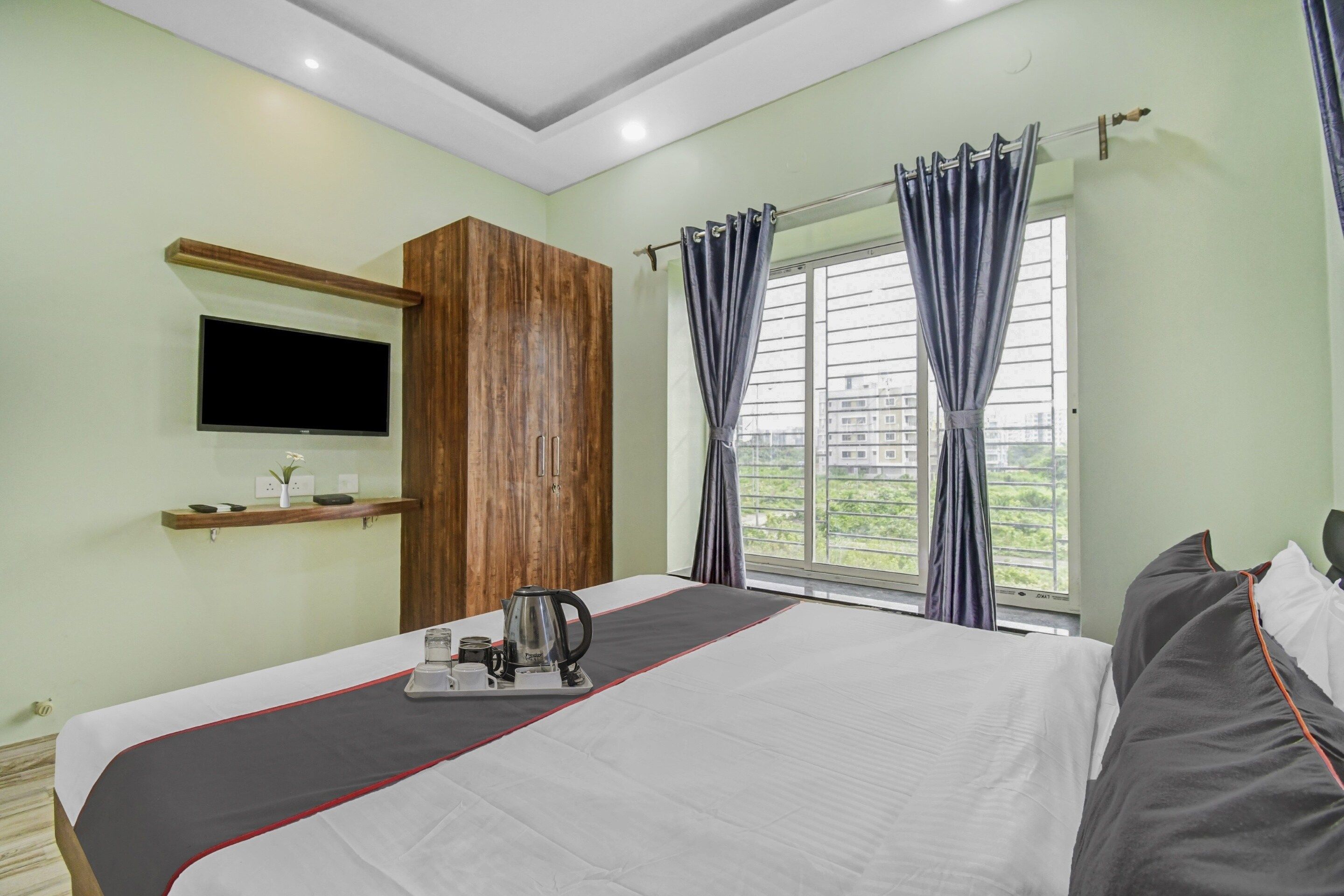 A hotel room with a bed, a TV mounted on the wall, a window with curtains, and a view of buildings outside.