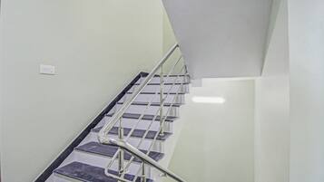 Una escalera con barandillas metálicas y peldaños oscuros, que lleva a un rellano con una pared blanca y un interruptor de luz.