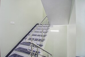 Eine Treppe mit Metallgeländern und dunklen Stufen, die zu einer Zwischenebene mit einer weißen Wand und einem Lichtschalter führt.