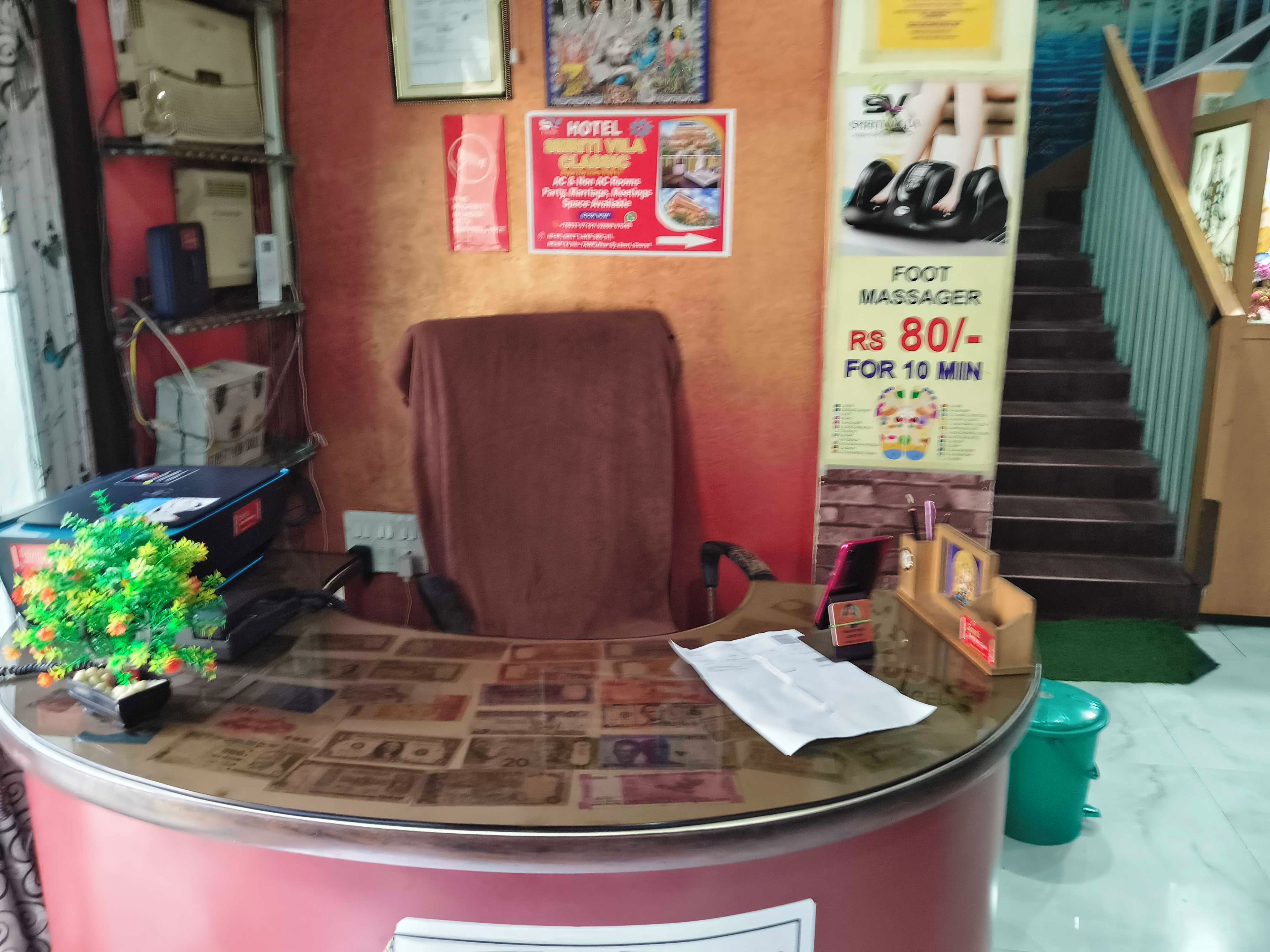 Un mostrador de recepción con una silla, una planta pequeña y un cartel publicitario de masajes podológicos.