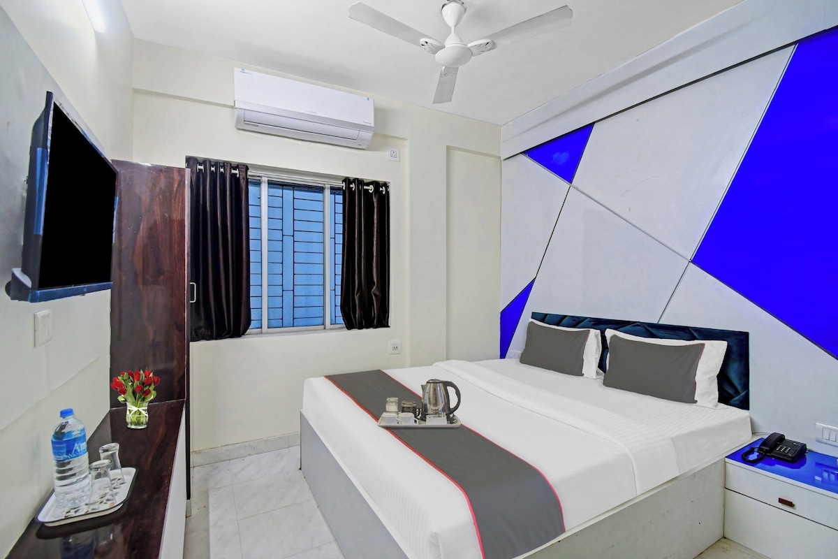 A hotel room with a bed, bedside table, a desk with a chair, a TV, an air conditioner, a window with curtains, and a ceiling fan.