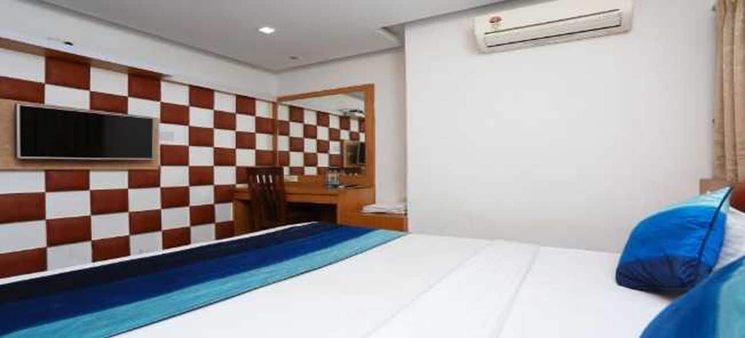 A hotel room with a checkered wall, a flat-screen TV mounted above a desk, a bed with a blue and white striped bedspread, and an air conditioning unit on the wall.