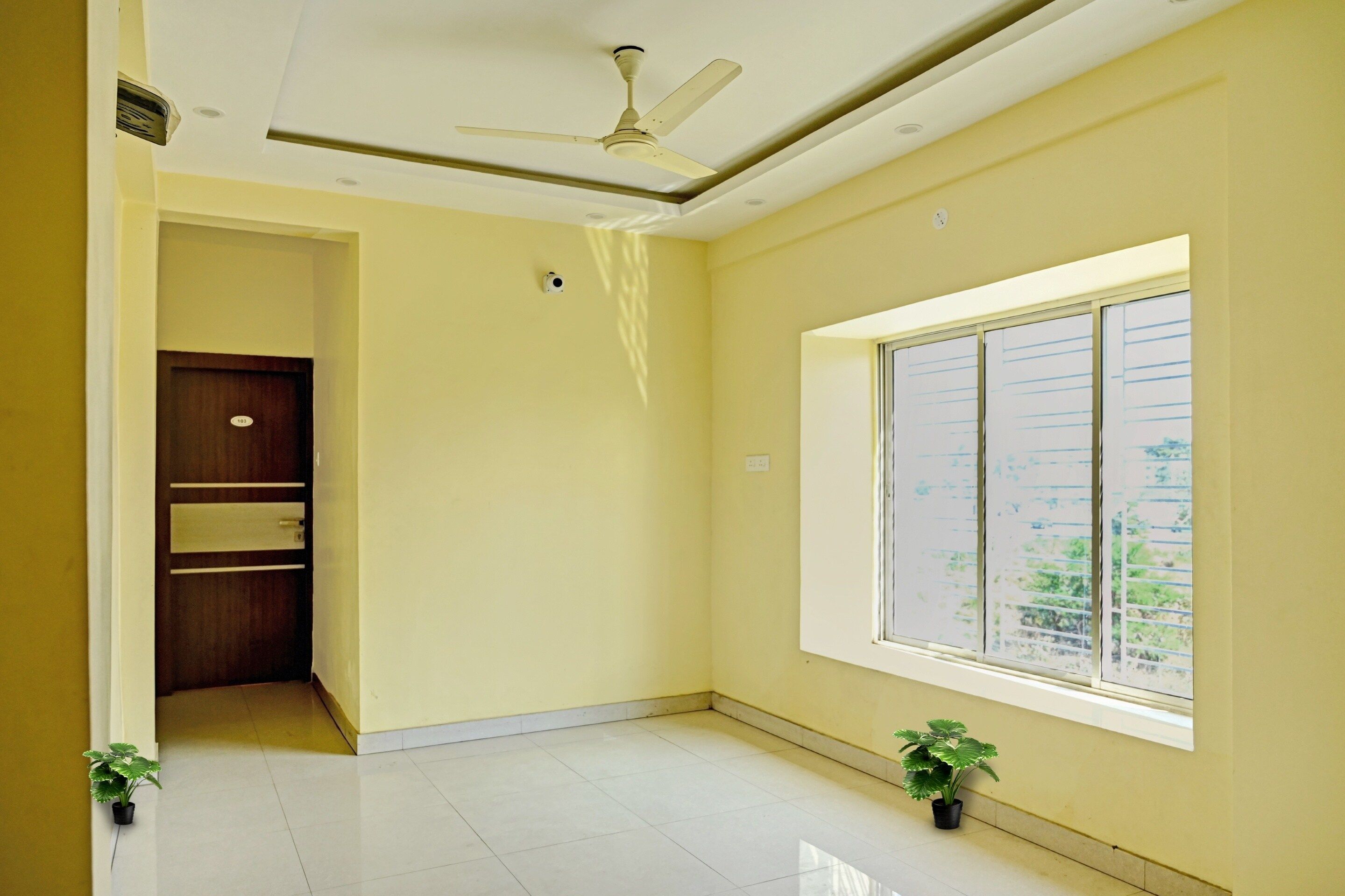 A room with a ceiling fan, a window with blinds, a door with a number, and two potted plants.
