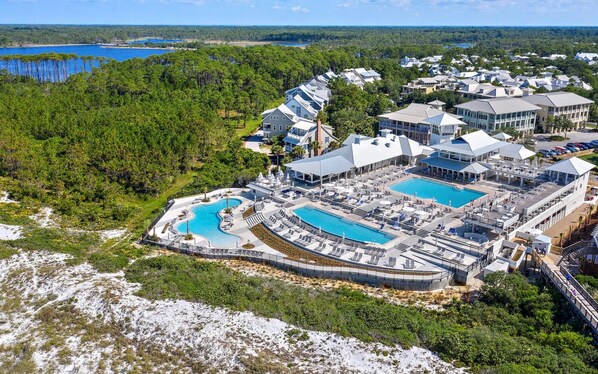 A resort with multiple swimming pools, a large building with a flat roof, and a forested area in the background. - WaterColor | 2 King Suites, Beach & Pools (Santa Rosa Beach)