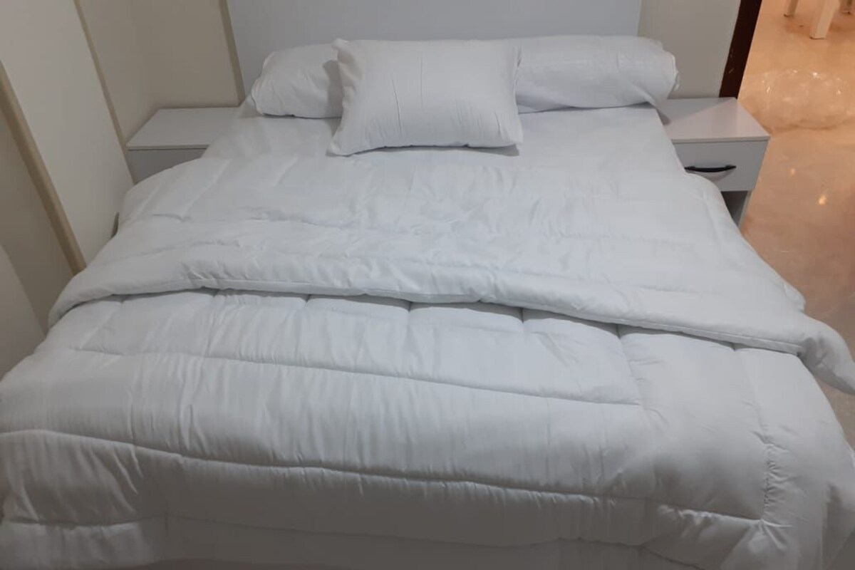 A bed with a white duvet and pillows, a nightstand with a drawer, and a tiled floor.