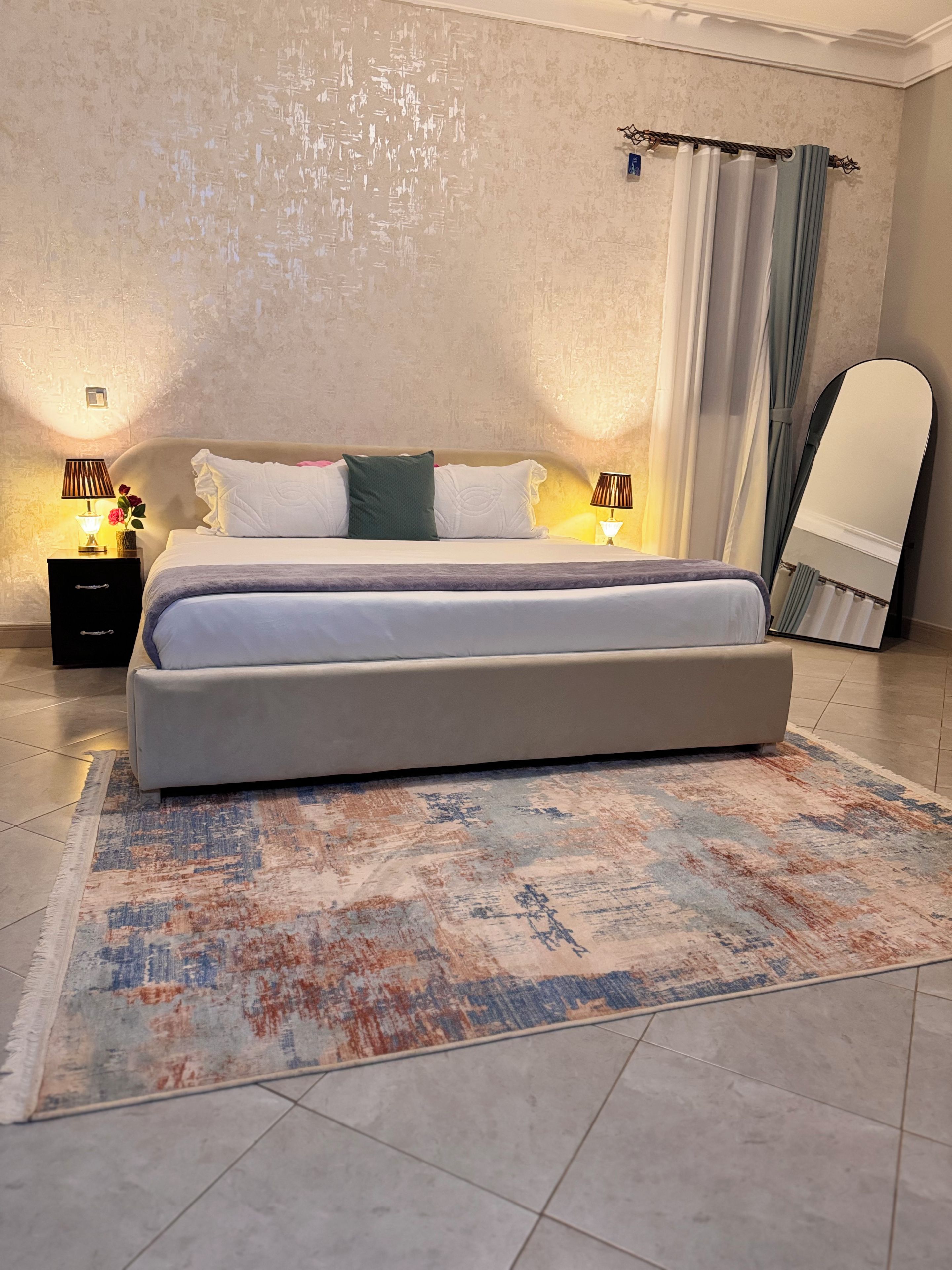 Een moderne slaapkamer met een groot bed, twee nachtkastjes met lampen, een decoratief vloerkleed en een lange spiegel.