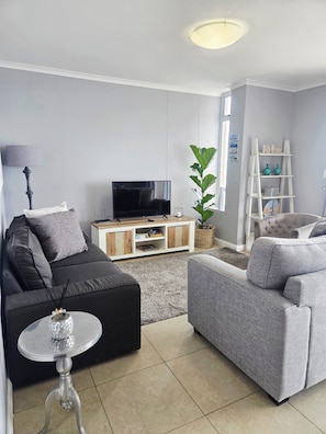 Un salon moderne avec un téléviseur à écran plat, un canapé gris, une petite table ronde et une plante en pot.