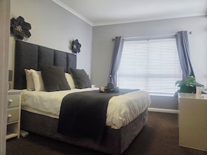 Ein Schlafzimmer mit einem großen Bett, einem Nachttisch, einer Kommode, einem Fenster mit Vorhängen und einer Wanddekoration.