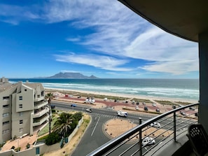 A view of a coastal road with vehicles, a beach, and a mountain in the distance. - Serene 2-bedroom apartment in Bloubergstrand perfect for your getaway (Cape Town)