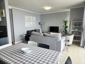 A living room with a sofa, a TV, a dining table with a checkered tablecloth, and a bookshelf. - Serene 2-bedroom apartment in Bloubergstrand perfect for your getaway (Cape Town)