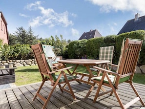 Eine Holzterrasse mit Tisch und Stühlen, umgeben von einer Hecke und einer Steinmauer.