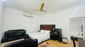 Una camera d'albergo con un letto, una scrivania, una sedia, una televisione, un condizionatore d'aria e una ventola a soffitto.
