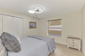 Ein Schlafzimmer mit einem Bett, einem Nachttisch, einem Fenster mit Jalousien, einer Deckenleuchte und einer Tür.