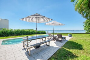A poolside area with a view of the water, a table, chairs, and umbrellas. - Sarasota Sunset Villa-Luxury waterfront pool home (Sarasota)