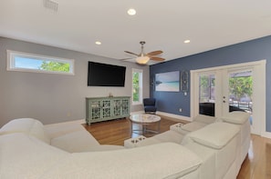 A living room with a ceiling fan, a flat-screen TV, a green cabinet, a blue armchair, and a glass coffee table. - Magnolia House Sarasota (Sarasota)