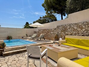 A pool area with a stone wall, lounge chairs, a yellow sofa, and a white chair. - Villa Sweet Nest with private heatable pool (Kassiopi)