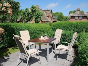 A patio with a table and four chairs, surrounded by well-maintained hedges and a thatched-roof building in the background. - Holiday Apartment in Wrixum With Terrace (Wrixum)