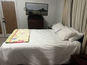 Ein Bett mit weißer Bettdecke und floralem Überwurf, ein Nachttisch mit Fernbedienung, ein an der Wand montierter Fernseher und ein Fenster mit Vorhängen.