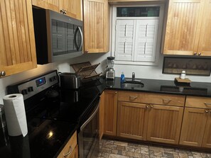 Una cocina con armarios de madera, encimera negra, microondas, tostadora y ventana con persianas.
