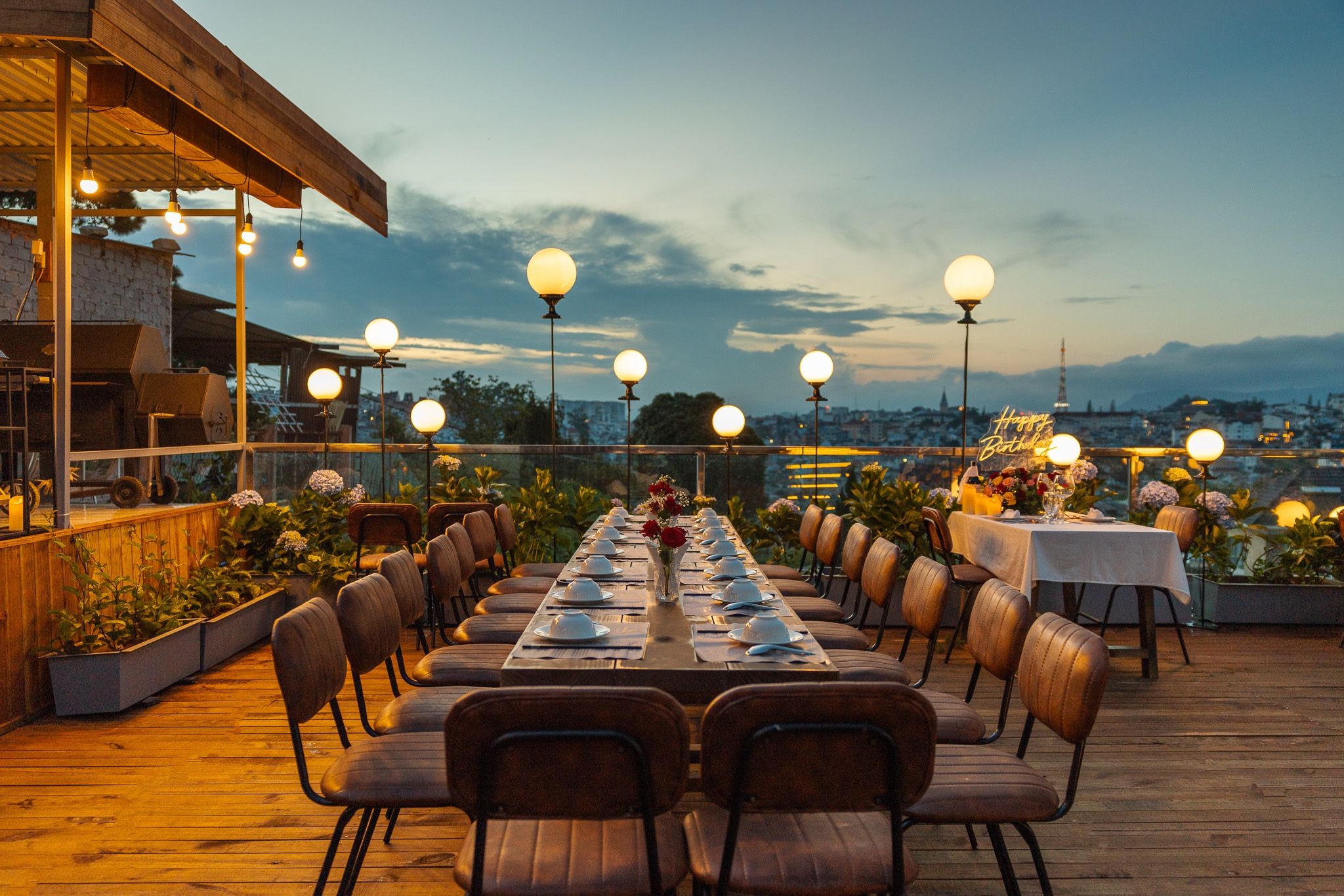 Un área de comedor al aire libre con una mesa larga dispuesta para una comida, rodeada de sillas e iluminada por luces esféricas.
