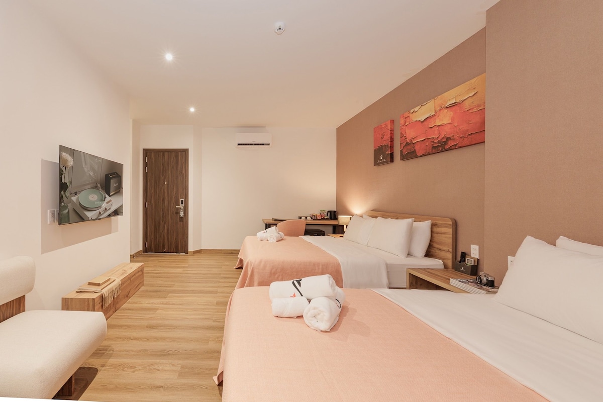 Een hotelkamer met een houten vloer, twee bedden met witte lakens, een houten hoofdeinde, een bureau met een lamp en kunst aan de muur.
