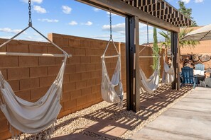 A series of white hammocks suspended from a wooden structure against a brick wall, with a patio area and outdoor seating visible in the background. - Prospector Peaks: Private Pool + Loads of Fun (Washington)