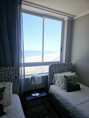 Een hotelkamer met twee bedden, een tafeltje, een lamp en een groot raam met uitzicht op het strand.