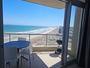 A balcony with a table and chairs, overlooking a beach and cityscape. - Perfect 2-bedroom apartment in Bloubergstrand for your getaway (Cape Town)