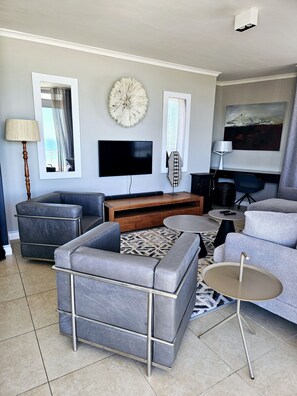 Een moderne woonkamer met een flatscreen televisie, grijze banken, een salontafel en een kamerkleed met een patroon.