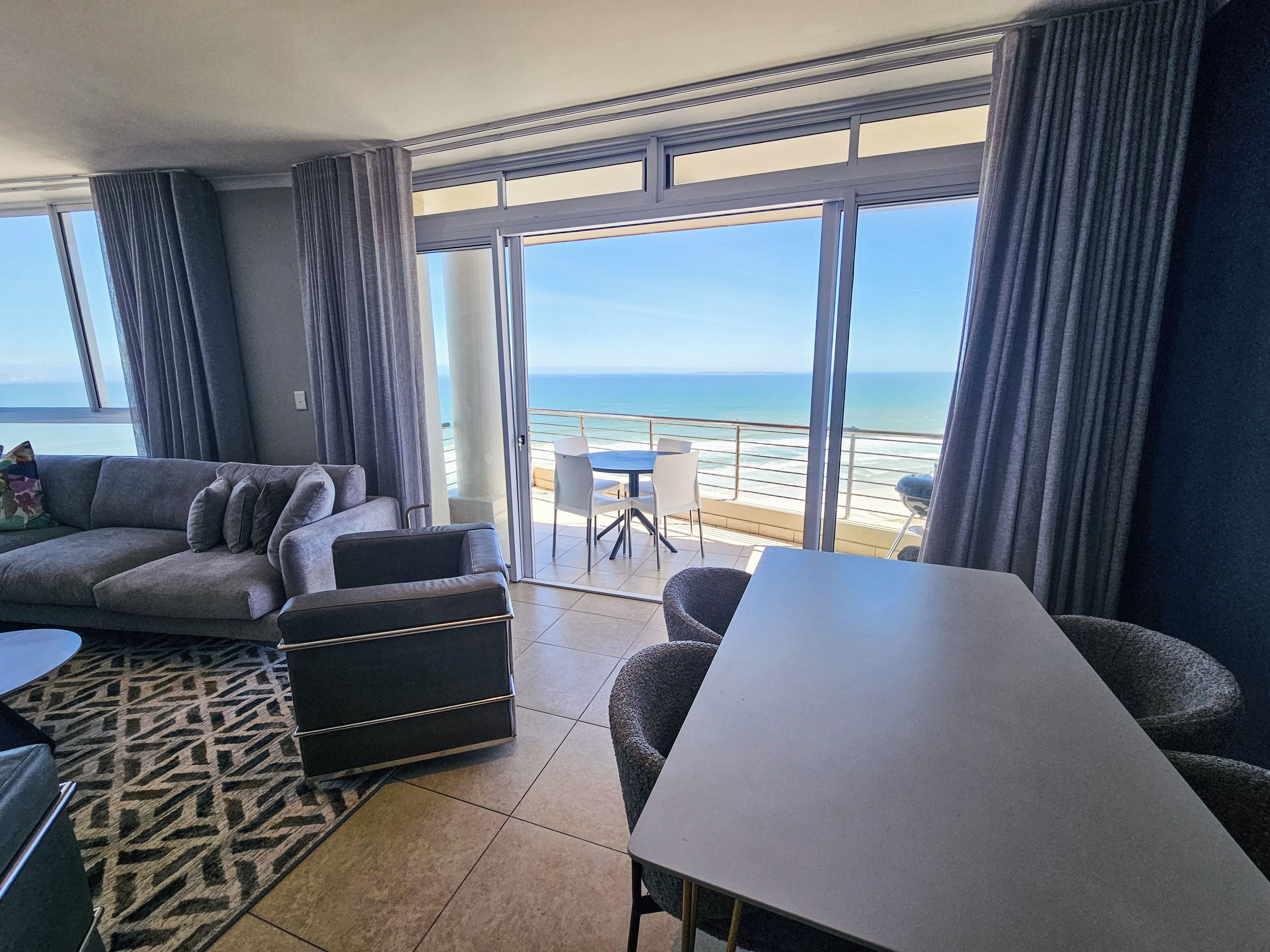 Ein modernes Wohnzimmer mit großem Fenster mit Blick auf einen Strand, einer Couch, einem Esstisch und einem gemusterten Teppich.