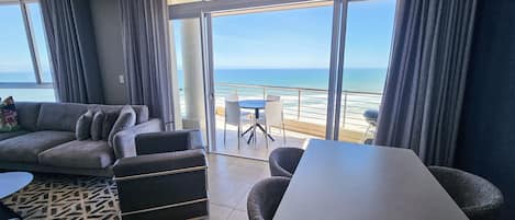 Un salon moderne doté d’une grande fenêtre donnant sur une plage, d’un canapé, d’une table à manger et d’un tapis à motifs.