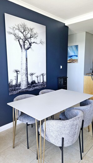 A dining area with a white table and chairs, a large tree painting on the wall, and a cabinet. - Perfect 2-bedroom apartment in Bloubergstrand for your getaway (Cape Town)