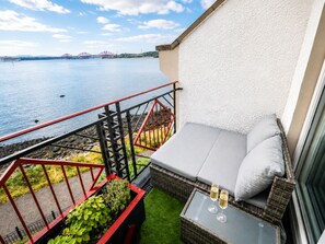 Een balkon met uitzicht op een brug over het water, een rieten bank met kussens en een glazen tafel met twee glaasjes.