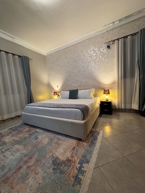 Quarto com uma cama grande, duas mesas de cabeceira com abajures, um tapete estampado e cortinas.