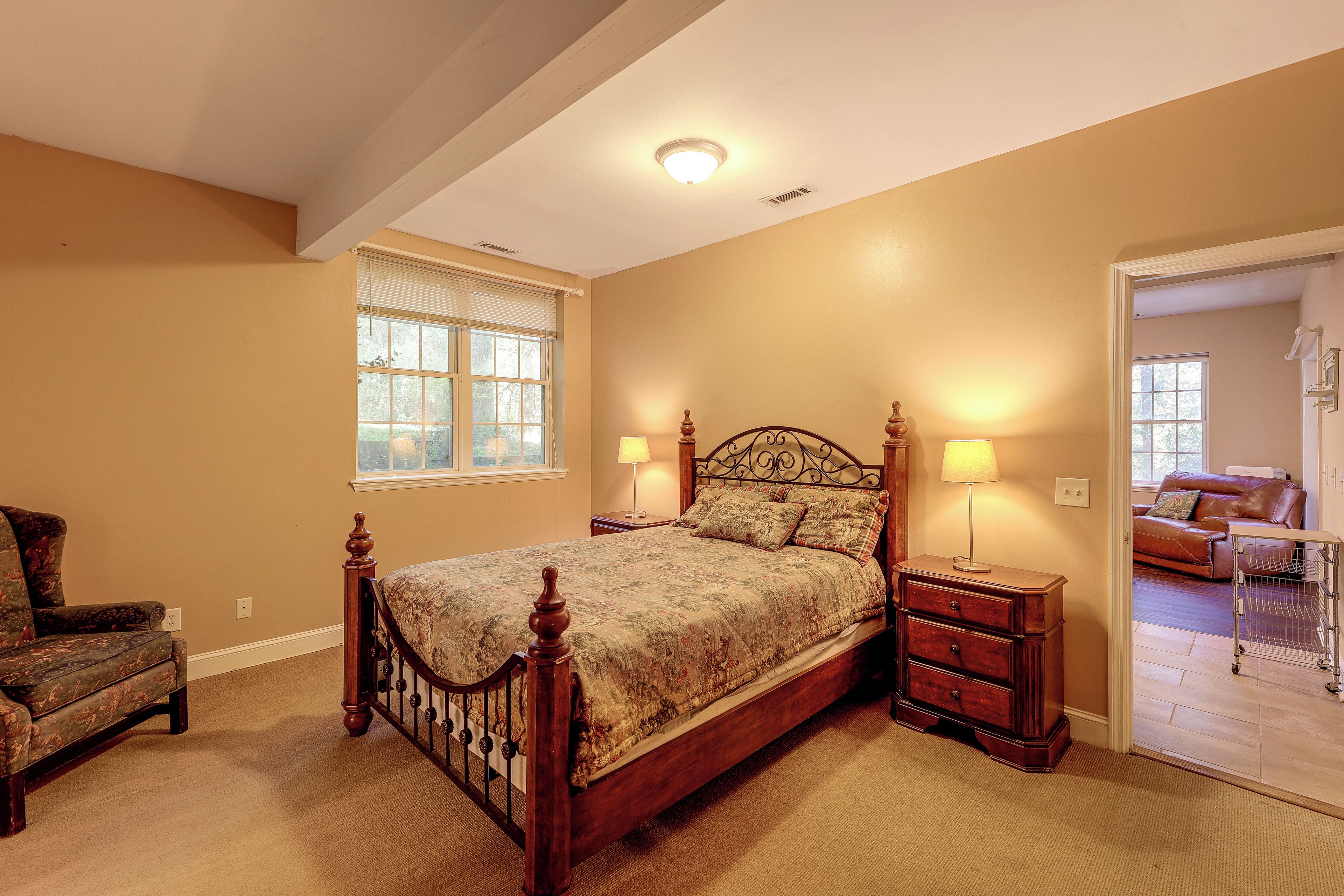 A bedroom with a wooden bed, two bedside lamps, a window with blinds, a chair, and a view of another room with a couch.