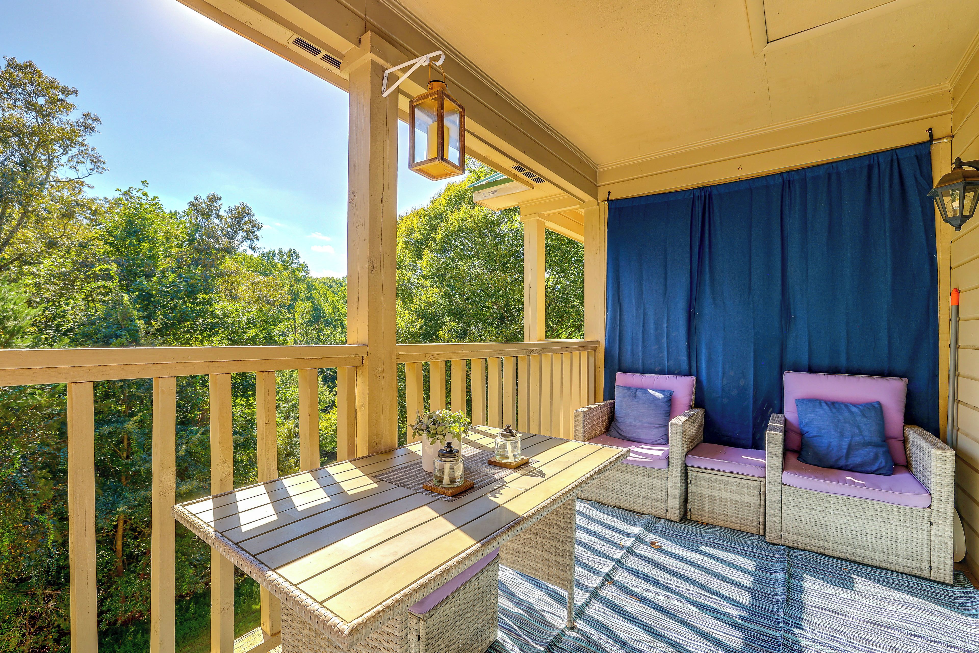 A wooden deck with a table and wicker furniture, a blue curtain, and a view of greenery.