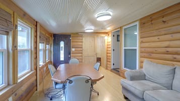 Interior de una cabaña de troncos con una mesa de comedor, sillas, un sofá y una puerta que da a otra habitación.