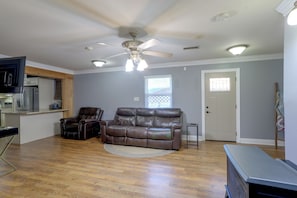 A living room with a brown leather sofa, a ceiling fan, and a kitchen area with appliances. - Bayou Days, Fire Pit Nights! Denham Springs Home (Denham Springs)