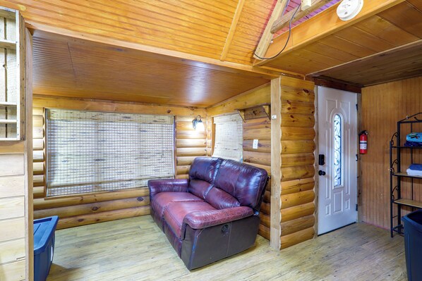 A log cabin interior with a leather sofa, a window with blinds, a door with a glass panel, and a shelf with items. - Private Porch & Yard: Denham Springs Cabin (Denham Springs)