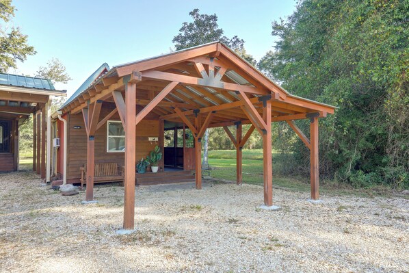 A wooden gazebo with a sloped roof, supported by wooden beams, situated on a gravel surface with a grassy area and trees in the background.