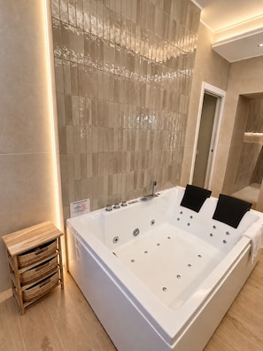 Een moderne badkamer met een groot wit bad, een houten bijzettafeltje en een betegelde muur met een uniek patroon.