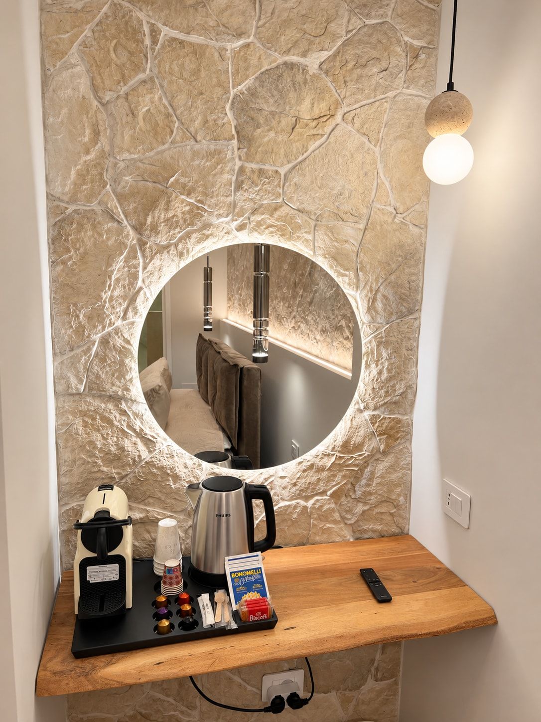 Un meuble-lavabo de salle de bain avec un mur en pierre, un miroir rond, une machine à café, une bouilloire, une télécommande et une petite étagère contenant des articles de toilette.