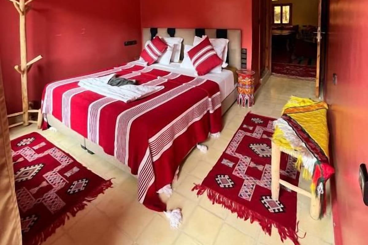 A room with a large bed, a bedside table, a chair, and a red wall.