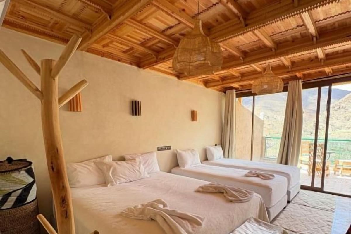 A room with two beds, a wooden ceiling, and a view of the outdoors.