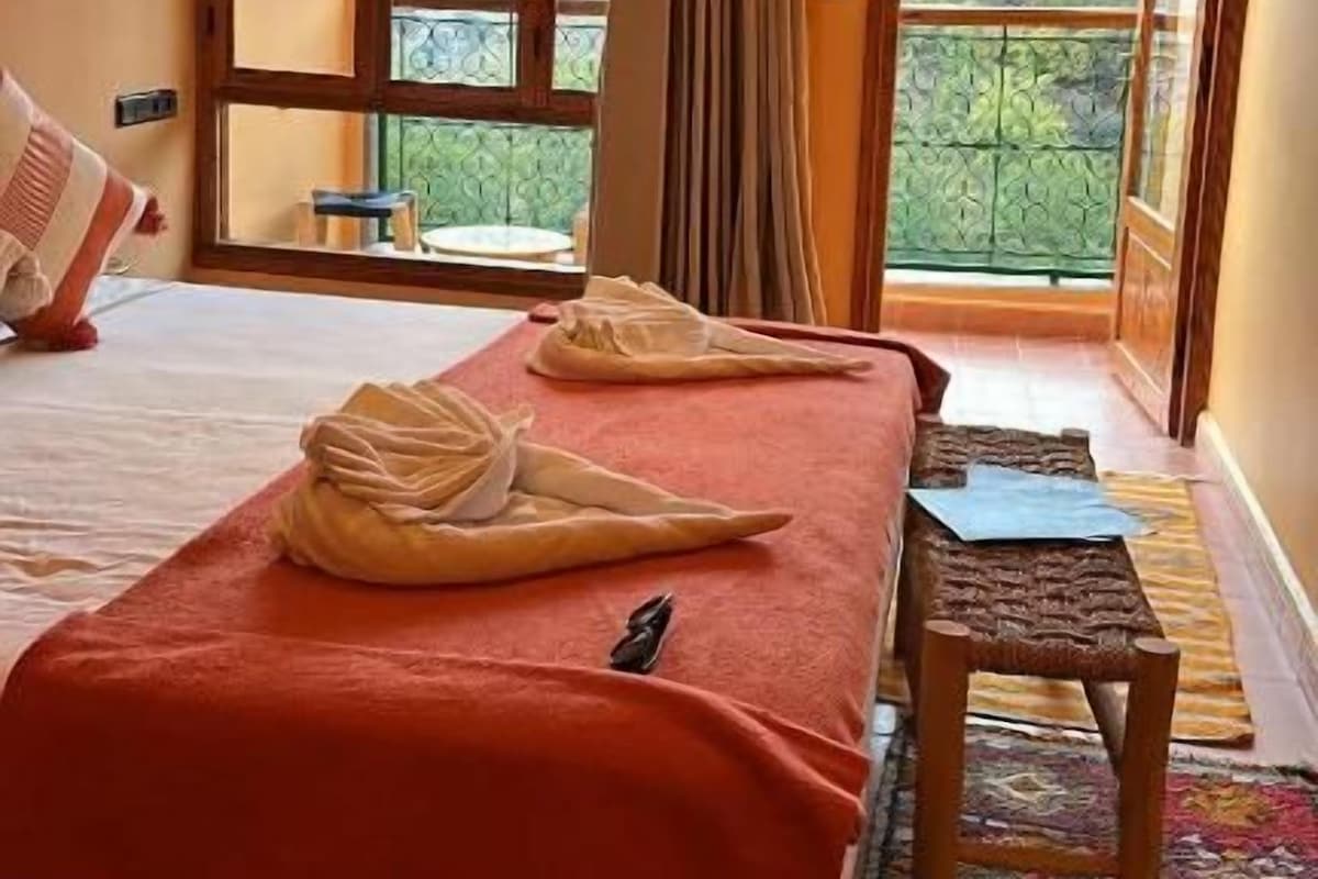A hotel room with a bed, a small table, a balcony with a view, and a hanging light fixture.
