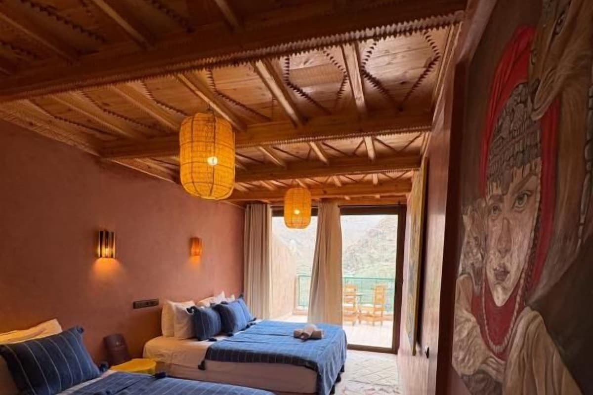 A hotel room with two beds, a wooden ceiling, a patterned rug, and a view of the outdoors.
