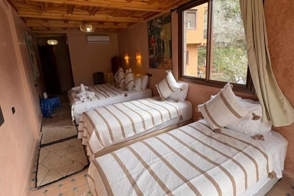 A room with a wooden ceiling, two beds with striped bedding, a window with curtains, and a door leading to another room.