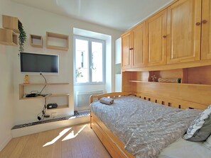 Un dormitori amb llit, armaris de fusta, televisió penjada a la paret i finestra que deixa entrar llum natural.