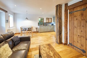 A spacious living area with a leather sofa, a wooden coffee table, and a dining table with chairs. A kitchen with grey cabinets and a wooden door is visible in the background. - Host Stay - Bricknell Barn (York)